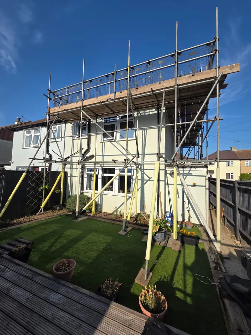 Rear elevation of the same house from the garden, scaffold running the full width with edge protection and boards set clear of the planting